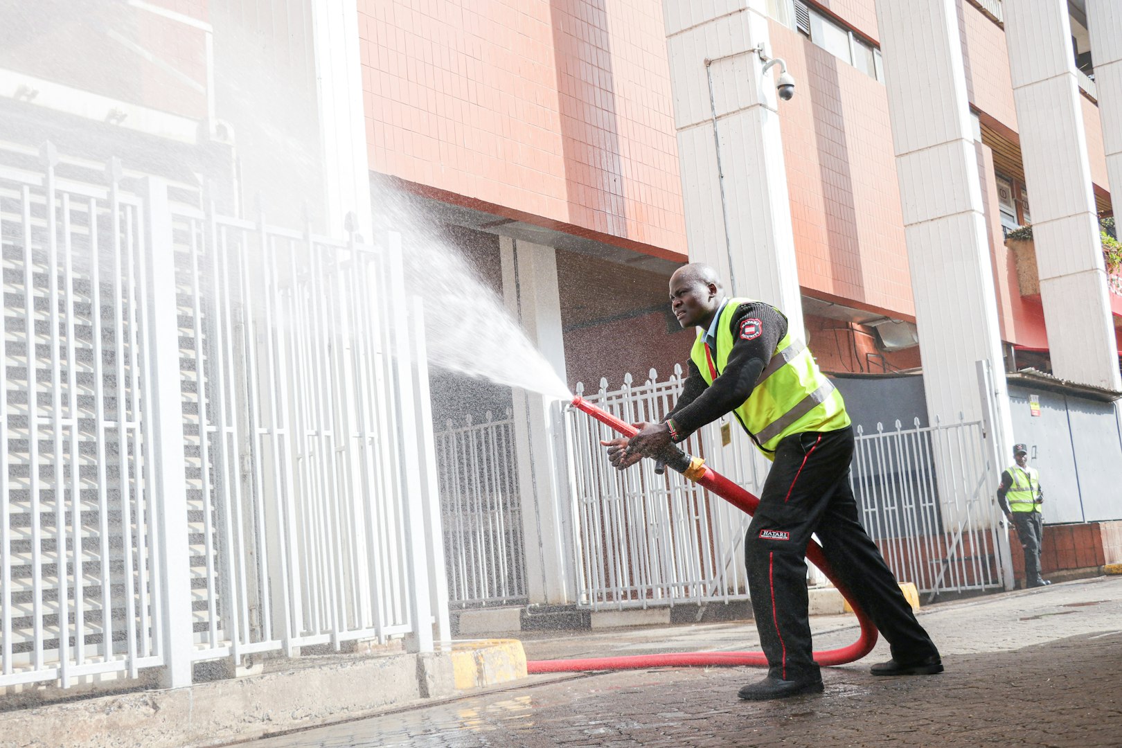 A person in overalls is using a pressure washer to clean a stone patio, with a lounge chair and neatly trimmed hedges visible in the background.